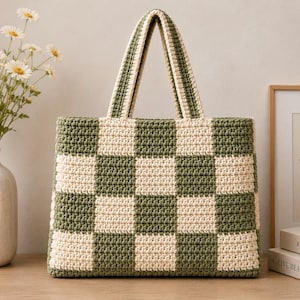 May include: A rectangular crochet tote bag featuring a repeating checkerboard pattern in olive green and cream yarn. The bag has two matching crocheted handles attached to the top edge, with a structured base and a consistent textured stitch throughout.