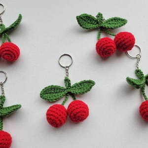 May include: Crocheted cherry keychains. Each keychain features two red cherries with green leaves and stems. The cherries are attached to a silver-colored key ring. The keychains are handmade and have a textured appearance.
