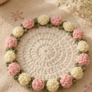 May include: A round, cream-colored crocheted coaster featuring a decorative border of small, three-dimensional pink and pale yellow yarn flowers interspersed with green crocheted leaves, creating a floral wreath pattern around the center.