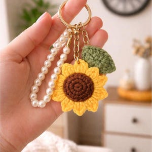May include: A handmade keychain featuring a yellow and brown crochet sunflower, a green crochet leaf, and a strand of white pearls. The keychain is attached to a gold-colored ring.