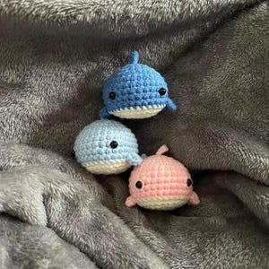 以下が含まれることがあります： 青とピンクの色合いの手作りの編みぐるみクジラのおもちゃ3つ。白いお腹と小さな黒い目があります。クジラは柔らかい灰色のテクスチャのある表面に配置されています。