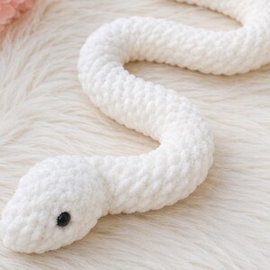May include: A long, white, crocheted snake soft toy with a textured, yarn-based surface. The snake features a rounded head with a single visible black, smooth, circular plastic eye. The body is coiled in a gentle S-shape, showcasing consistent, uniform stitches throughout the entire length of the stuffed animal.
