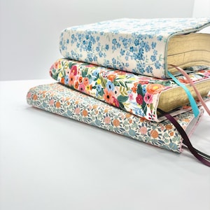 Può includere: Una pila di tre libri con copertine a motivi floreali. Il libro superiore ha un design floreale blu chiaro, il libro centrale ha un motivo floreale colorato e il libro inferiore ha un motivo floreale più piccolo con accenti arancioni e blu. Ogni libro ha un segnalibro a nastro.