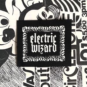 Può includere: Toppa quadrata nera con la scritta "electric wizard" in bianco, in carattere gotico, incorniciata da un bordo decorativo bianco. La toppa è posta su uno sfondo in bianco e nero con vari elementi grafici.
