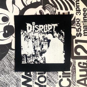 Può includere: Una toppa quadrata nera con la parola "DISRUPT" in un carattere gotico distorto sopra una grafica bianca di una figura e un paesaggio urbano. La toppa è posta su uno sfondo di manifesti in bianco e nero.