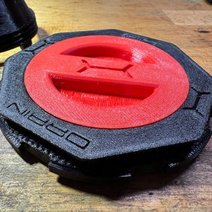 以下が含まれることがあります： 側面に「ORBIN」の文字が入った、黒と赤の3Dプリントオブジェクト。赤い円形の中央と隆起したデザインを持つ六角形です。木製の表面に置かれています。