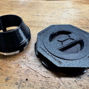 May include: Black 3D-printed automotive parts, including a circular collar and a hexagonal cap with the text "OIL CATCH" and "SANTA" visible. The parts are made of a textured material and sit on a wooden surface.