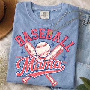 Peut inclure: T-shirt bleu clair avec l'inscription "BASEBALL Mama" en rouge et blanc. Le motif comprend une balle de baseball, des battes croisées et des étoiles. Le t-shirt est plié et présenté avec un collier et un jean.