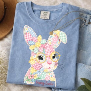 Puede incluir: Camiseta Comfort Colors azul claro con un diseño de conejito patchwork. El conejito tiene gafas y una corona de flores, hecha de tela estampada rosa, amarilla y azul. El texto "Happy Easter" está debajo.