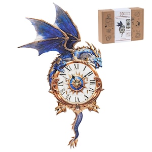 May include: A decorative clock featuring a blue dragon with outstretched wings perched atop a clock face with Roman numerals. The clock has a gold-colored frame and a blue tail. A cardboard box with the words "3D PUZZLE" is in the background.