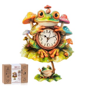 May include: A 3D wooden puzzle clock featuring a large green frog with orange accents sitting on a lily pad, surrounded by colorful mushrooms. A smaller frog sits on a lower lily pad base. The clock face has black numbers and hands. Includes a cardboard box with the text 3D WOODEN PUZZLE.