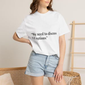 May include: White t-shirt with the text "We need to discuss VE options" in black. The shirt is paired with denim shorts. A light-colored wooden ladder and a woven basket are in the background.