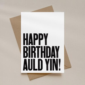 May include: A white greeting card with the text "HAPPY BIRTHDAY AULD YIN!" in bold black letters. The card is placed on top of a brown envelope, with a neutral background.