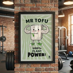 Può includere: Un poster incorniciato con un personaggio di tofu dei cartoni animati con le braccia muscolose, scarpe da ginnastica verdi e una foglia. Il poster recita "MR TOFU" e "100% PLANT POWER!" su uno sfondo verde chiaro. Il poster è esposto in una palestra.