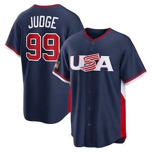 May include: Navy blue baseball jersey with red and white accents. The front features the word "USA" in white with red stripes, and the back displays "JUDGE" and the number "99" in red. The jersey has a button-up front and a small American flag patch.