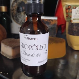 May include: A brown glass spray bottle with a black cap and a white label. The label reads "Jicote Prop&oacute;leo Para la tos". The bottle is filled with a light brown liquid. The background includes other products.