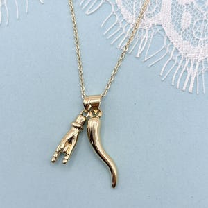 May include: A gold-toned necklace featuring two charms suspended from a delicate chain. One charm is a hand making a rock-and-roll gesture, and the other charm is a curved, tapered horn shape. Both charms have a polished, reflective metallic finish.