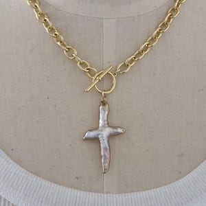 May include: Gold-tone chain necklace with a cross pendant. The necklace features a chunky chain design and a toggle clasp. The cross pendant has a pearl-like appearance with gold-tone edging. The necklace is displayed on a mannequin.
