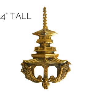 May include: A gold-colored metal pagoda-style wall decor with intricate details. It is 14 inches tall and features a dragon-like design on the bottom.