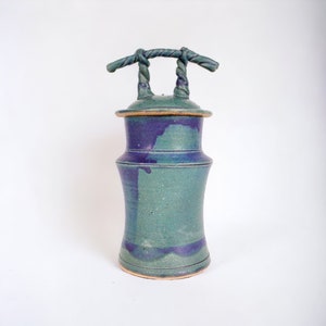 May include: A green and purple ceramic jar with a lid and a twisted rope handle. The jar has a speckled glaze and a unique shape.