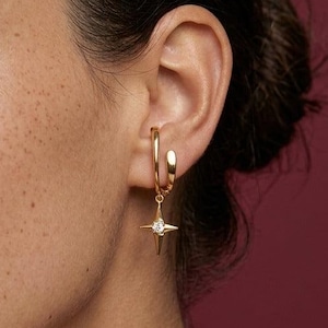 Könnte beinhalten: Nahaufnahme einer Person, die einen goldenen Creolen-Ohrring mit einem Stern-Anhänger trägt. Der Ohrring hat einen kleinen, klaren Edelstein in der Mitte des Sterns. Der Hintergrund ist ein dunkler Rotton.