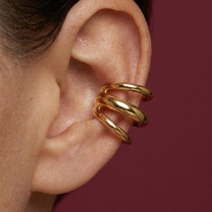 Puede incluir: Un ear cuff dorado con un diseño en espiral. El ear cuff se envuelve alrededor de la oreja con tres bandas conectadas. La joya es brillante y reflectante, y el fondo es de color rojo oscuro.