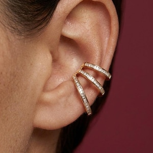 Puede incluir: Puño de oreja dorado con tres bandas apiladas, cada una adornada con pequeñas piedras brillantes. La joya se lleva en la oreja, sobre un fondo burdeos. Un accesorio de oreja moderno y elegante.