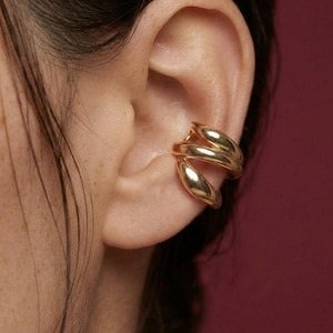 Könnte beinhalten: Ein goldener Ohrklemme umschließt das Ohr. Der Ohrschmuck hat eine glatte, polierte Oberfläche und ein dreifaches Schlaufendesign. Der Schmuck wird vor einem weinroten Hintergrund gezeigt.