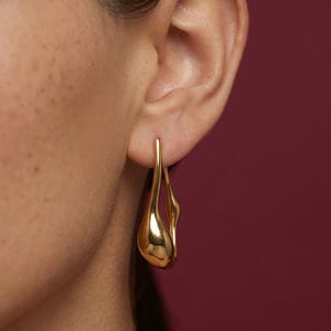 Könnte beinhalten: Nahaufnahme eines goldfarbenen Ohrrings. Der Ohrring hat eine einzigartige, organische Form mit einer glatten, reflektierenden Oberfläche. Das Design scheint eine Träne oder eine fließende Form zu sein, mit poliertem Finish.