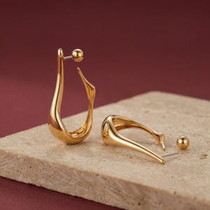 May include: A pair of gold-colored, abstract-shaped earrings. One earring is fully assembled, while the other is partially disassembled, with a small ball stud separate. The earrings are displayed on a light-colored, textured surface against a burgundy background.