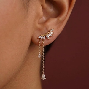 May include: A gold ear cuff featuring a cluster of clear marquise-cut stones and two dangling teardrop-shaped stones. The ear cuff is worn on the ear and has a delicate chain design.