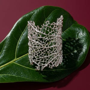 May include: A silver cuff bracelet with a detailed, openwork design, similar to coral or a web. The bracelet is presented on a large, glossy green leaf, set against a deep red backdrop.