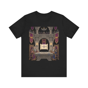 May include: Black t-shirt with a detailed illustration of a medieval council scene. Two robed figures sit at a table, with text on a scroll. The background includes an arched structure, banners, and additional figures.