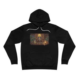 Peut inclure: Sweat-shirt à capuche noir avec une illustration détaillée d'un magicien dans une pièce avec des écrans d'ordinateur. Le magicien est assis à un bureau avec une lumière vive au-dessus de lui. Le sweat-shirt a une poche avant et un cordon blanc.