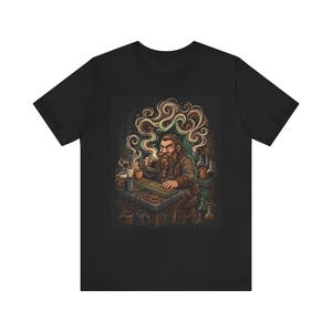 May include: Black t-shirt with a fantasy-themed illustration. It depicts a bearded character with a mug, seated at a desk with a keyboard and mugs. The artwork includes swirling steam and laboratory flasks.