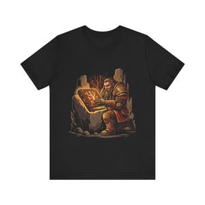 Op de afbeelding: Zwart T-shirt met een kleurrijke illustratie van een bebaarde figuur in harnas, knielend voor een gloeiend boek met printplaatontwerpen. Het kunstwerk is geplaatst tegen een rotsachtige achtergrond, met een warme, verlichte gloed.