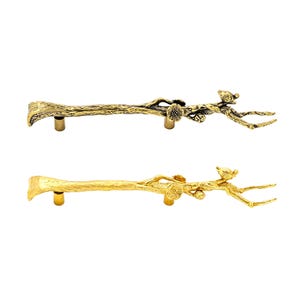 May include: Two metal cabinet pulls shaped like tree branches with textured bark, small flowers, and a bird perched on the end. One pull has an antique dark brass finish, while the other has a bright, polished gold finish, each with two mounting posts.