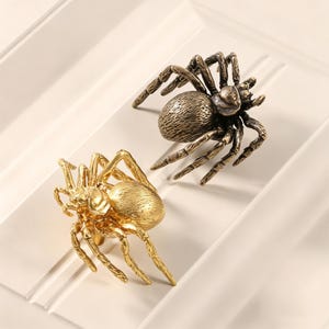 Puede incluir: Dos pomos para armario con forma de araña, uno con acabado en tono dorado brillante y el otro en un tono bronce antiguo más oscuro. Cada pomo presenta un cuerpo detallado y texturizado con ocho patas articuladas que se extienden hacia fuera.