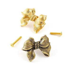 May include: Two bow-shaped cabinet knobs with a textured, ribbed surface and a central beaded band. One knob features a bright gold finish, while the other has an antiqued brass finish. Two matching gold-toned metal mounting screws are included.