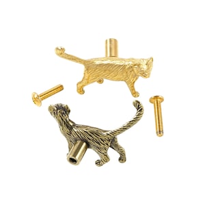 May include: Two cat-shaped cabinet knobs made of polished brass, each featuring a textured feline silhouette with a raised tail. The set includes two matching metal screws for installation, with one knob shown from the side and one from the back.