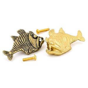 May include: Two fish-shaped cabinet knobs with detailed scales and fins. One knob has an antique brass finish with dark highlights, while the other has a bright, polished gold finish. Two matching gold-toned metal screws are included for installation.