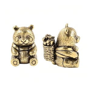 May include: Two brass panda figurines with a metallic gold finish. One panda is shown from the front, while the other is shown from the side, featuring a detailed woven basket strapped to its back filled with small cylindrical bamboo stalks.