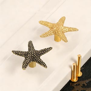 May include: Two starfish-shaped cabinet knobs, one in a bright gold finish and one in a dark bronze finish, each featuring a textured, bumpy surface and a cylindrical base. Two threaded metal mounting screws are included for installation.