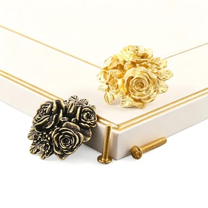 May include: Two metal cabinet knobs shaped like clusters of roses, one in a bright gold finish and one in an antique bronze finish. Each knob features detailed floral petals and leaves, accompanied by two matching threaded metal mounting screws.