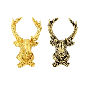 Könnte beinhalten: Zwei Hirschkopf-Anstecknadeln aus Metall mit Geweih. Eine Nadel hat ein helles, poliertes Gold-Finish, während die andere ein antikes, dunkleres Messing-Finish aufweist. Beide zeigen ein detailliertes Tiergesicht und ein strukturiertes Halstuch.