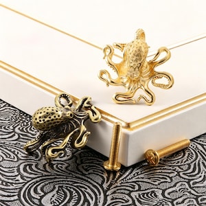 Puede incluir: Dos pomos para armario con forma de pulpo en acabados de oro metálico y latón antiguo. Cada pomo presenta un manto texturizado y tentáculos curvados. Se incluyen dos tornillos de metal de color dorado a juego para su instalación.