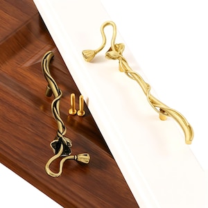 May include: Two metal cabinet pulls featuring a curved, vine-like design with a floral bud at one end. One pull has a polished gold finish, while the other has an antique bronze finish. Two small gold-colored mounting screws are included.