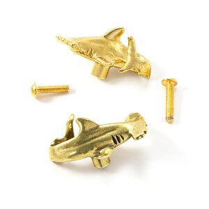 Könnte beinhalten: Zwei goldfarbene Schrankknöpfe aus Metall in Form von Hammerhaien, jeweils mit detaillierter Rückenflosse und Schwanz. Das Set enthält zwei passende goldfarbene Metallschrauben mit abgerundeten Köpfen zur Montage an Möbeln.