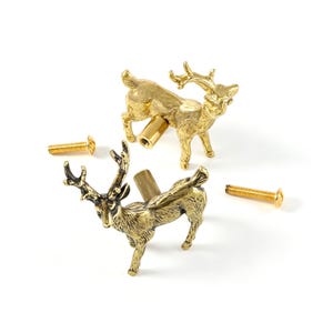 May include: Two gold-toned metal cabinet knobs shaped like standing deer with detailed antlers and bodies. Each knob features a cylindrical mounting post on the back and comes with a separate matching gold-toned metal screw for installation.