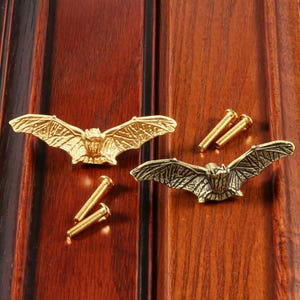 Puede incluir: Dos pomos para armario con forma de murciélago y acabado metálico, uno en tono dorado brillante y otro en tono latón antiguo, cada uno acompañado de un tornillo metálico roscado a juego para su instalación en muebles.